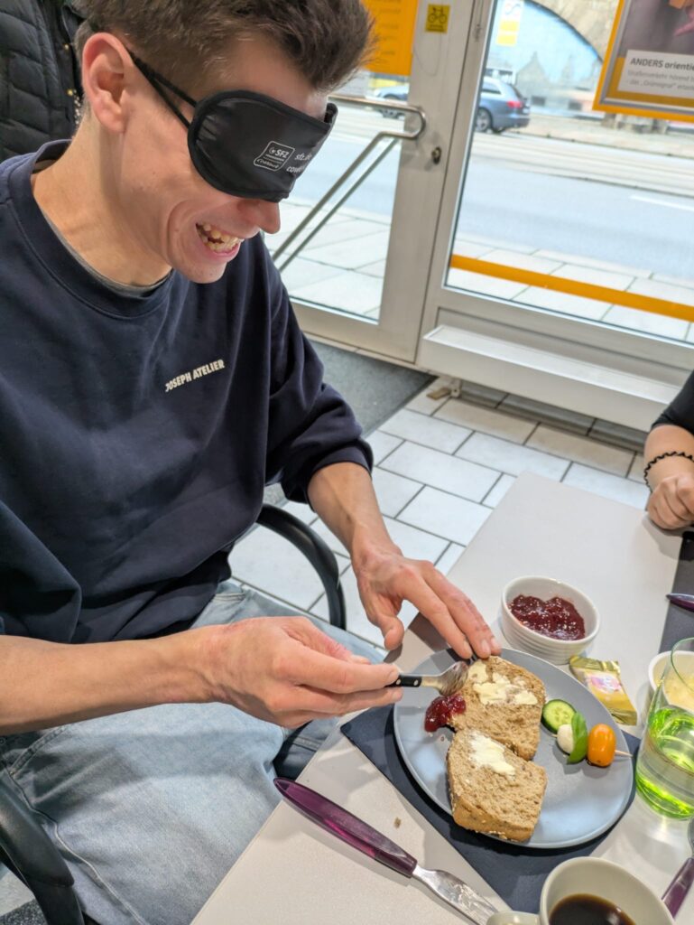 Das Bild zeigt einen Teilnehmer des Sensibilisierungskurses, er trägt eine Augenbinde und sitzt an einem Tisch. Vor ihm steht ein Frühstücksgedeck und auf seinem Teller liegen ein Gemüsespieß und ein Brötchen, das er mit Butter und Marmelade bestreichen möchte. Die Butter und Marmelade sind nur teilweise auf dem Brötchen verteilt, ein Teil befindet sich daneben auf dem Teller. Der Teilnehmer lacht, weil ihm ein Missgeschick passiert ist.