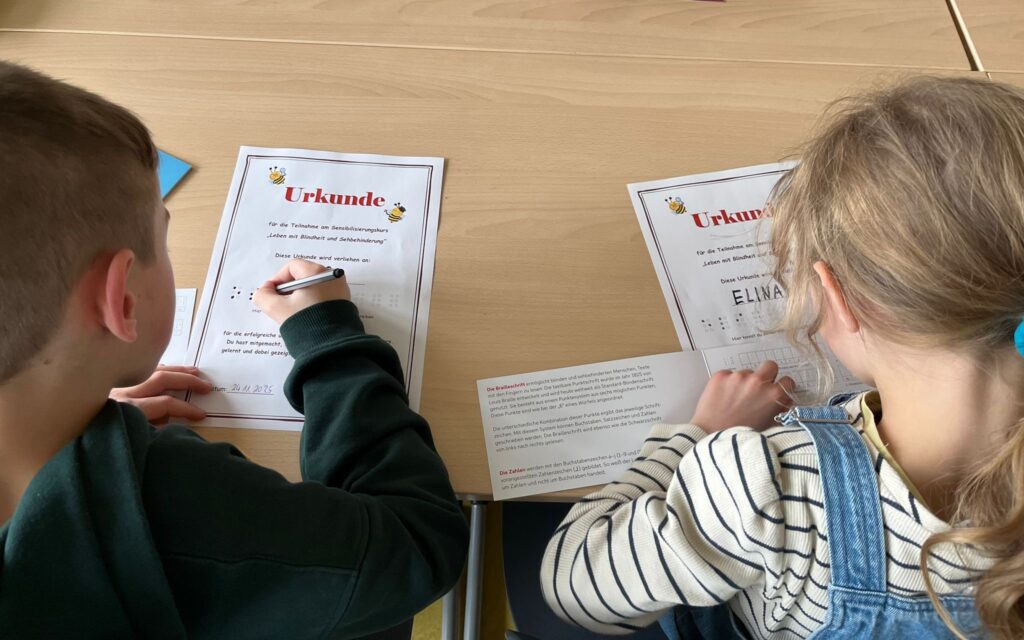 Das Bild zeigt zwei Kinder einer 3. Klasse, von hinten fotografiert, die an einem Tisch sitzen. Vor ihnen liegen Urkunden, die sie für die Teilnahme am Sensibilisierungskurs ‚Leben mit Blindheit und Sehbehinderung‘ erhalten haben. Die Kinder schreiben ihren Namen in Brailleschrift in Brailleschrift-Zeilen auf die Urkunden. Neben den Urkunden liegt ein Brailleschrift-Alphabet, das als Vorlage dient.