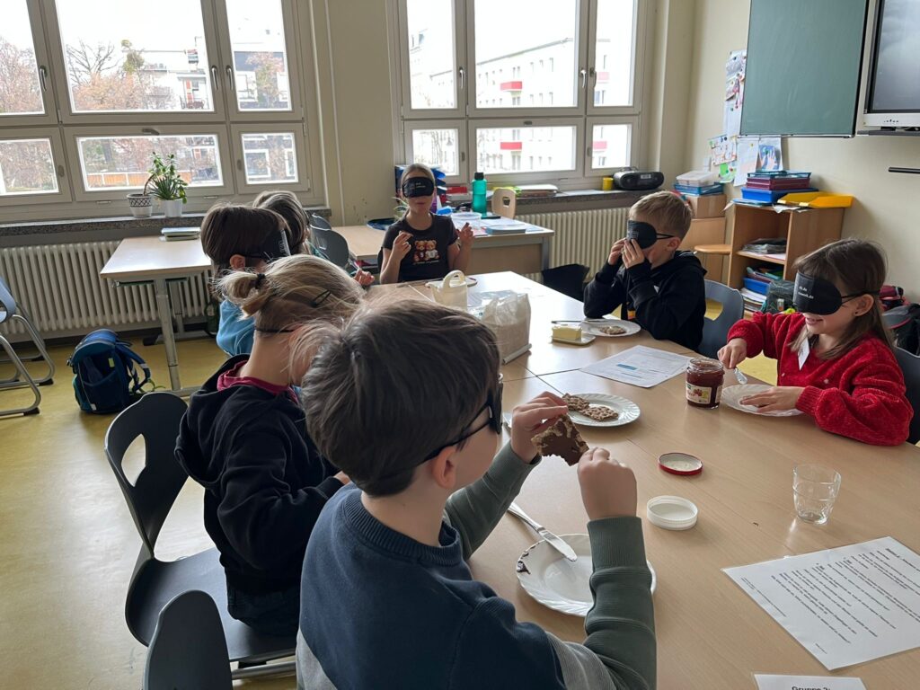 Das Bild zeigt mehrere Schulkinder einer 3. Klasse, die in einem Klassenraum an einem großen Tisch mit Frühstücksutensilien sitzen. Sie tragen Augenbinden. Einige schmieren noch Brote mit Butter und Marmelade, andere essen bereits ihre selbst geschmierten Brote. Manche Kinder halten ihre Hände hoch, die teilweise mit Marmelade und Butter verschmiert sind.