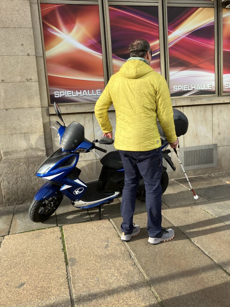 Das Bild zeigt eine Person, die von hinten zu sehen ist. Diese Person geht mit einem weißen Blindenstock auf einem Gehweg. Die Person tastet sich vorsichtig an einem blauen Moped vorbei, das am Rand des Gehweges steht