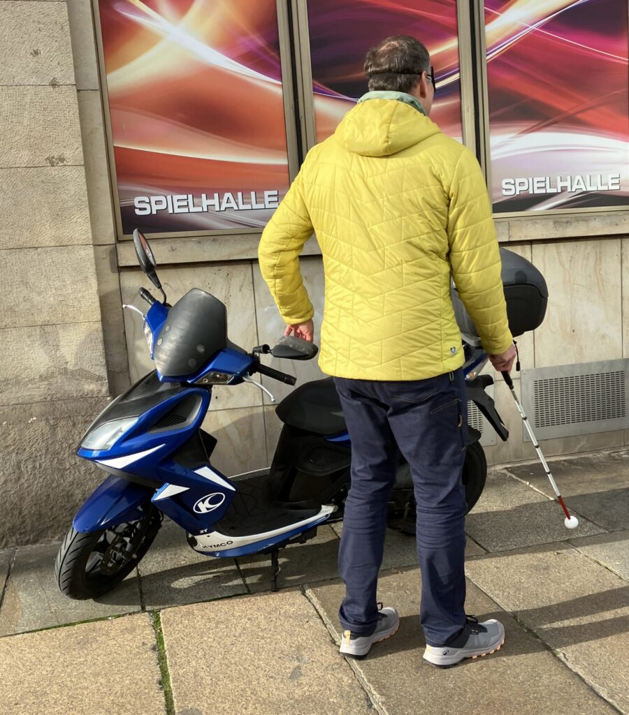 Das Bild zeigt eine Person, die von hinten zu sehen ist. Diese Person geht mit einem weißen Blindenstock auf einem Gehweg. Die Person tastet sich vorsichtig an einem blauen Moped vorbei, das am Rand des Gehweges steht