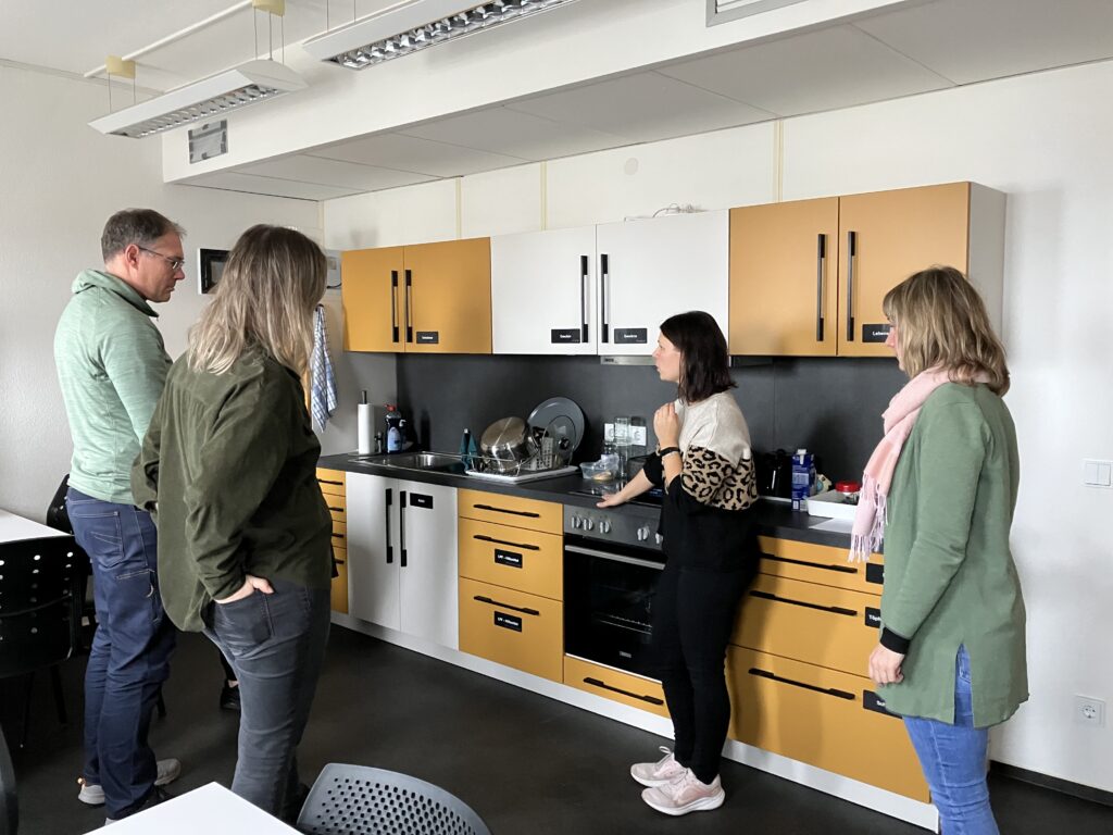 Das Bild zeigt einen Raum mit einer Küche, die deutliche Kontraste in Weiß, Dunkelgelb und Schwarz aufweist. Schränke und Geräte sind mit blindentechnischen Beschriftungen versehen. In der Mitte steht eine Person, die blindentechnische Hilfsmittel für den Haushalt erklärt. Daneben stehen drei weitere Personen, Teilnehmende eines Sensibilisierungskurses, die aufmerksam zuhören und sich etwas zeigen lassen.