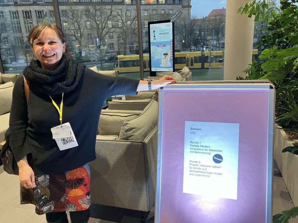 Das Bild zeigt eine Mitarbeiterin des SFZ Dresden, die auf der Konferenz Inklusion vor Ort in Dresden das Projekt Inklusion Sehen präsentiert. Sie steht in einem modernen Foyer des neuen Stadtforums Dresden neben einem beleuchteten Infoständer, auf dem ein Blatt mit Projektdetails angebracht ist. Im Hintergrund sind große Glasfenster mit Blick auf Straßenbahnen und das alte Dresdner Rathaus zu sehen.
