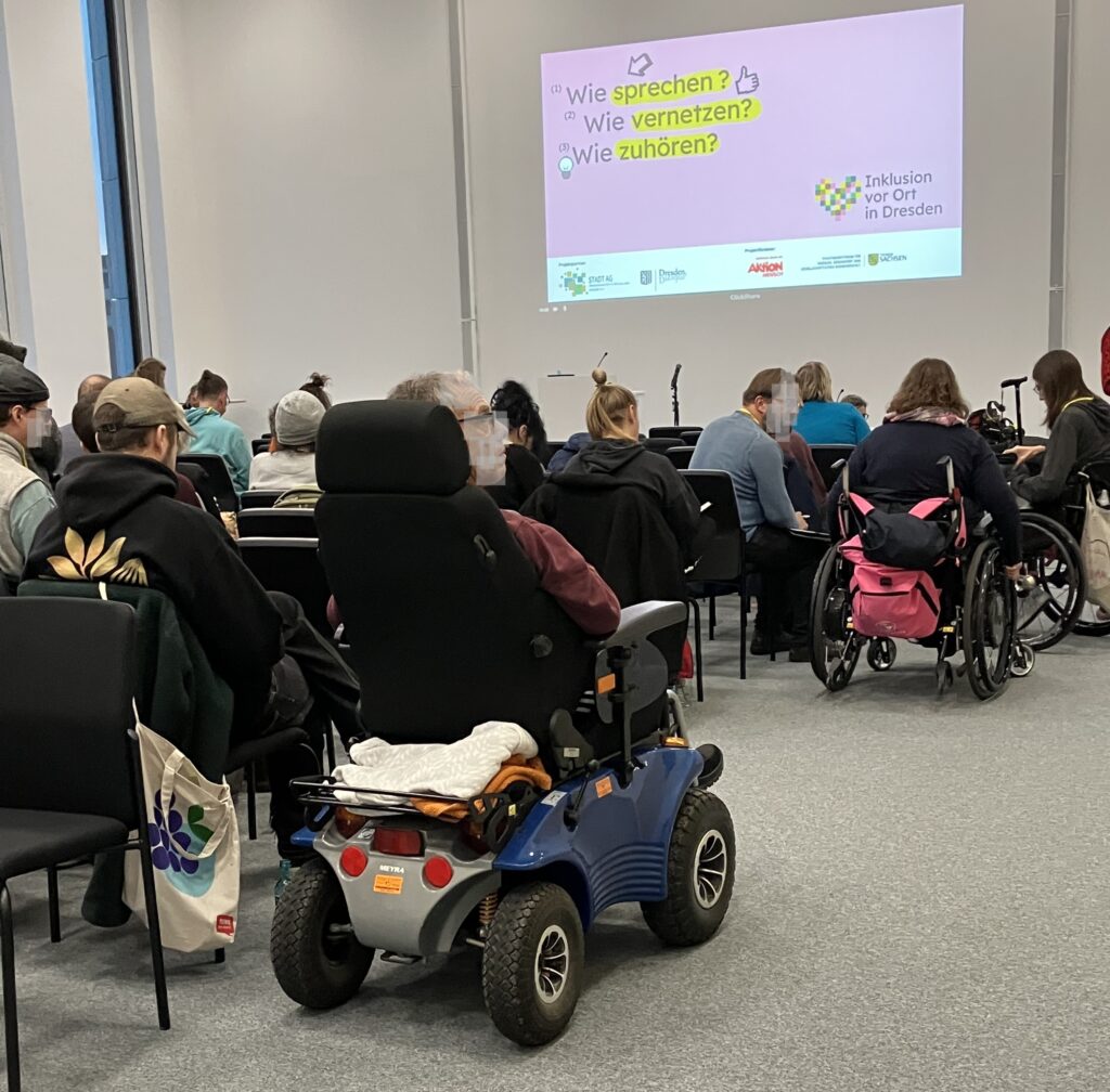 Auf dem Bild sieht man einen Konferenzsaal, der von hinten fotografiert ist. Mehrere Personen sitzen in Reihen, darunter auch mehrere Rollstuhlfahrer. Vorne ist eine große Projektionsfläche zu sehen, auf der eine Folie mit dem Titel ‚Inklusion vor Ort in Dresden‘ angezeigt wird. Die Folie enthält drei Themenfragen: Wie sprechen?, Wie vernetzen? und Wie zuhören?.