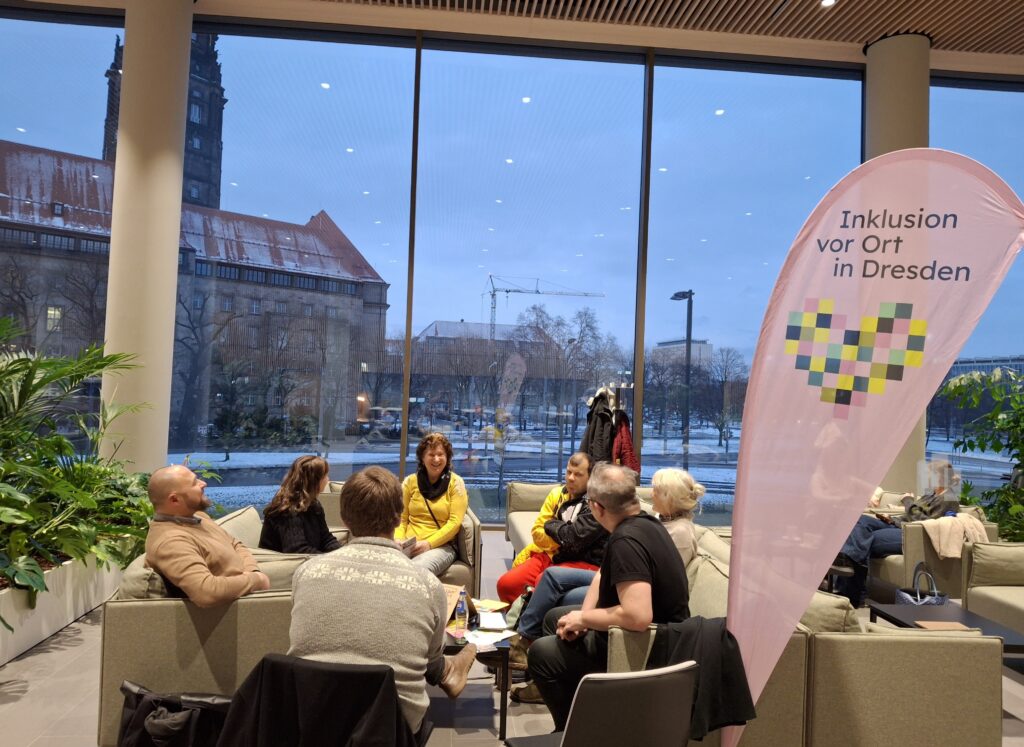Das Bild zeigt eine Gesprächsrunde im Stadtforum Dresden. Durch die großen Fenster im Hintergrund ist das Dresdner Rathaus zu sehen. Mehrere Personen sitzen in bequemen Sesseln und auf Couches zusammen und unterhalten sich in entspannter Atmosphäre. In der Mitte steht ein Tisch mit ausliegenden Flyern. Neben der Sitzgruppe befindet sich ein großes Segelbanner mit der Aufschrift „Inklusion vor Ort in Dresden“.