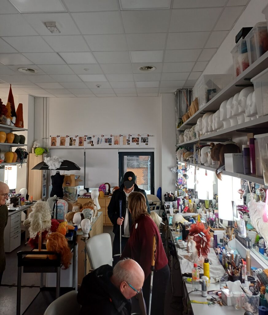 Das Bild zeigt einen hellen Raum der Theatermaske mit mehreren nebeneinanderliegenden Schminkplätzen. Auf den Tischen stehen und liegen zahlreiche Pinsel, Dosen, Fläschchen und andere Schminkutensilien. Über jedem Platz hängen große Spiegel, umrahmt von runden leuchtenden Lampen. Im Raum befinden sich viele unterschiedliche, auch verrückte, Perücken auf Styroporköpfen in verschiedenen Höhen, außerdem Hüte und weitere Requisiten auf Regalen und Arbeitstischen. Die Szenerie wirkt lebendig und leicht unaufgeräumt, so, als würde gerade kreativ gearbeitet werden.