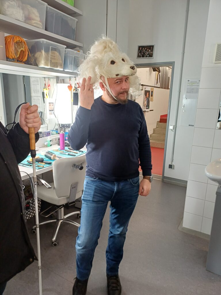 Das Bild zeigt einen unserer Mitarbeitenden, der eine weiße Pudelperücke anprobiert. Er steht dabei fröhlich in der Theatermaske und wirkt sichtlich gut gelaunt, während er mit der ausgefallenen Perücke Spaß hat.