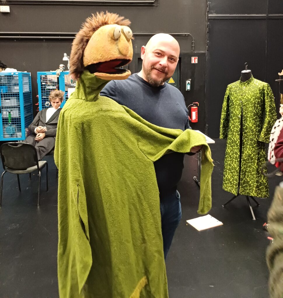 Das Bild zeigt einen unserer Mitarbeitenden, der eine große, witzige Handpuppe in Menschengröße lebhaft bespielt. Er hält die Puppe vor sich und bewegt sie so, als würde er sich mit ihr unterhalten. Die Szene spielt in der Requisite und wirkt fröhlich und verspielt, da der Mitarbeitende sichtlich Freude beim Umgang mit der Puppe hat.