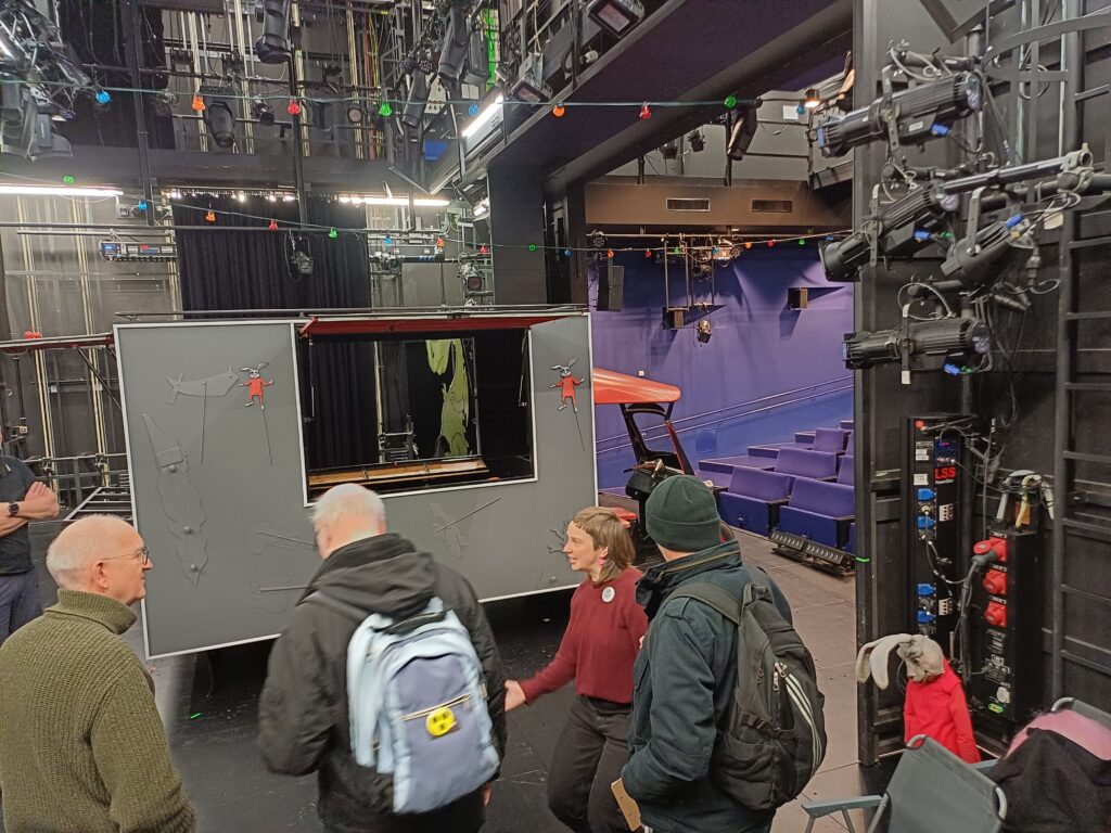 Das Bild zeigt die Rückseite eines Bühnenbildes mit sichtbarer Bühnentechnik wie ein umgebautes Tuk-tuk, Requisiten, Metallstreben, Beleuchtungsketten und Scheinwerfern. Zwischen den technischen Elementen steht eine Mitarbeiterin des tjg. und erklärt einer Gruppe von Teilnehmenden, wie sich das Bühnenbild bewegen und verändern lässt.